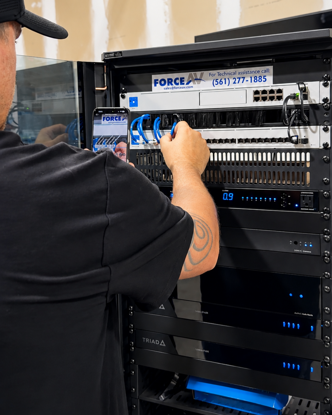 A technician in a black shirt working on a tall server rack, connecting a blue ethernet cable.