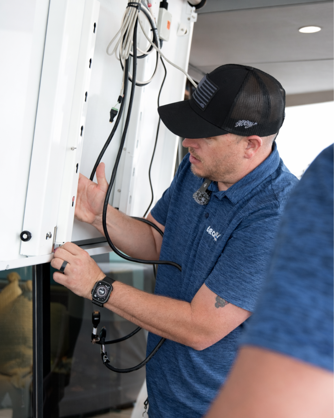 A man wearing a black baseball cap and blue shirt inspecting wires behind a white wall panel or enclosure.