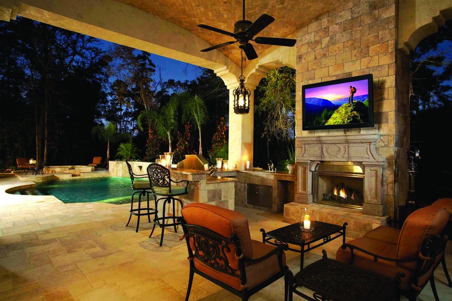 An outdoor patio with a Seura TV above a fireplace, an outdoor kitchen area, and a pool.